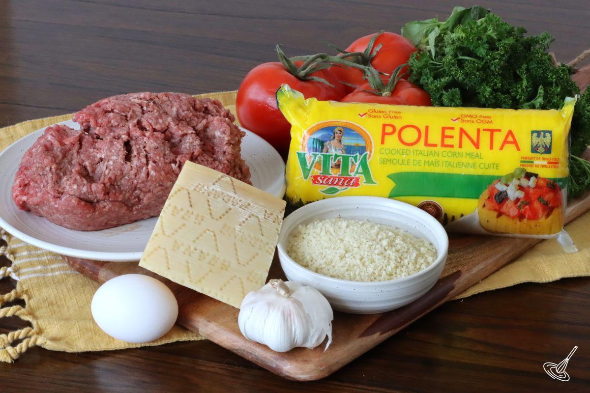 Ingredients on a wooden board including; panko crumbs, ground beef, egg, polenta and tomatoes.