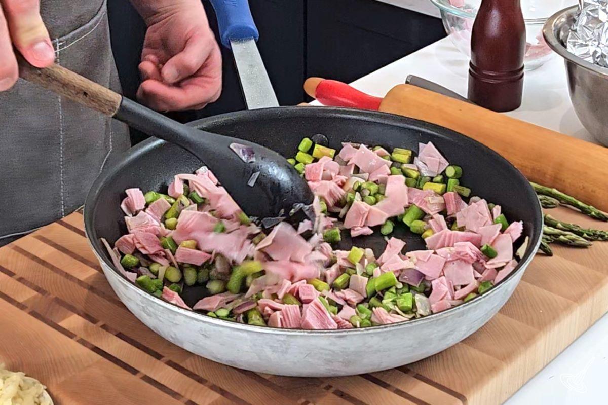 Quelqu'un qui fait revenir du jambon haché dans une grande poêle avec des oignons hachés et des asperges.