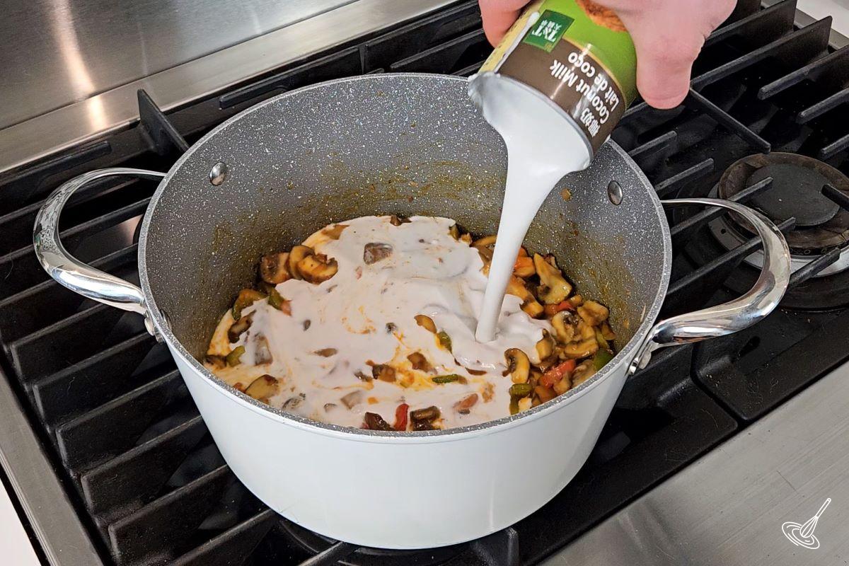 Someone adding coconut milk to a soup pot with red curry vegetables.