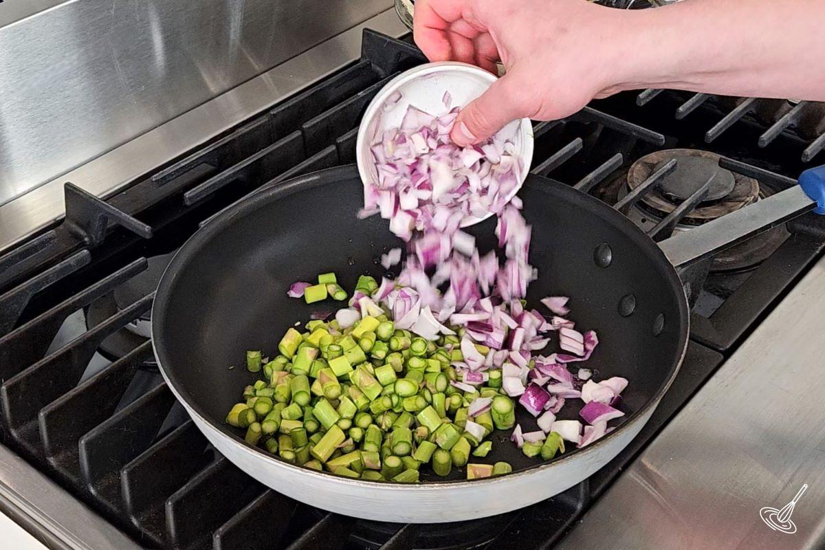 Quelqu'un qui place des oignons hachés dans une grande poêle avec des asperges hachées.