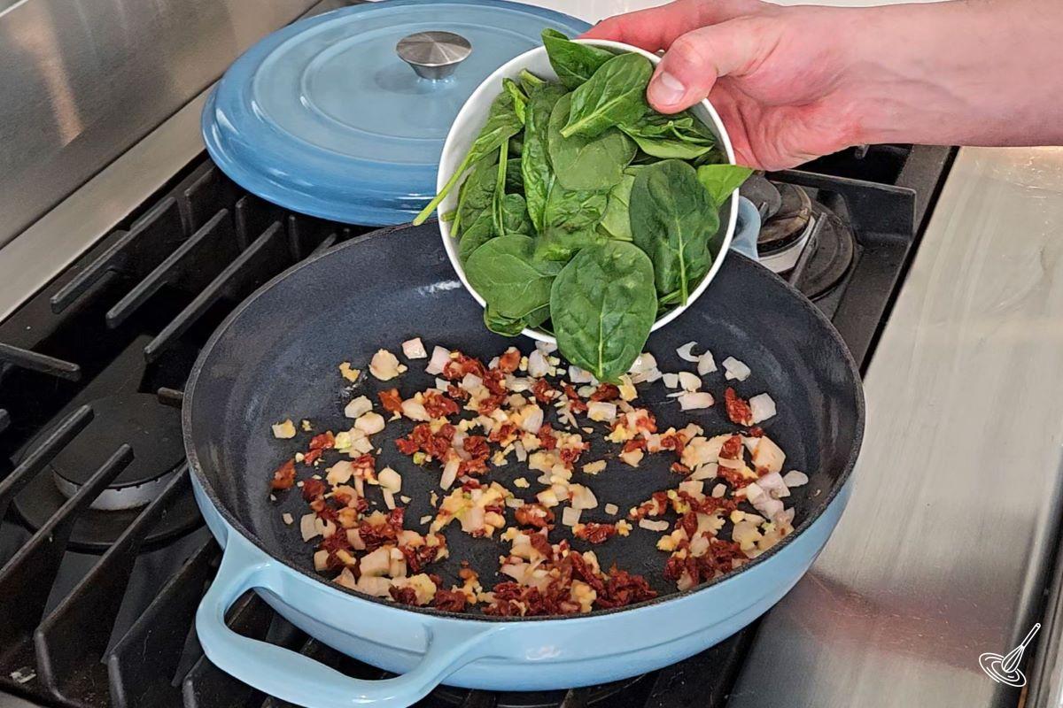 Someone adding baby spinach to a skillet with sundried tomatoes and shallot.