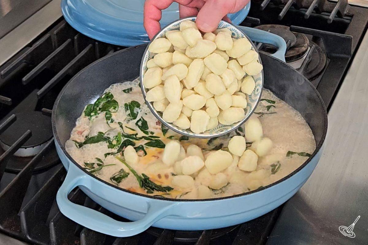 Someone adding gnocchi to a large skillet containing a cream sauce. 
