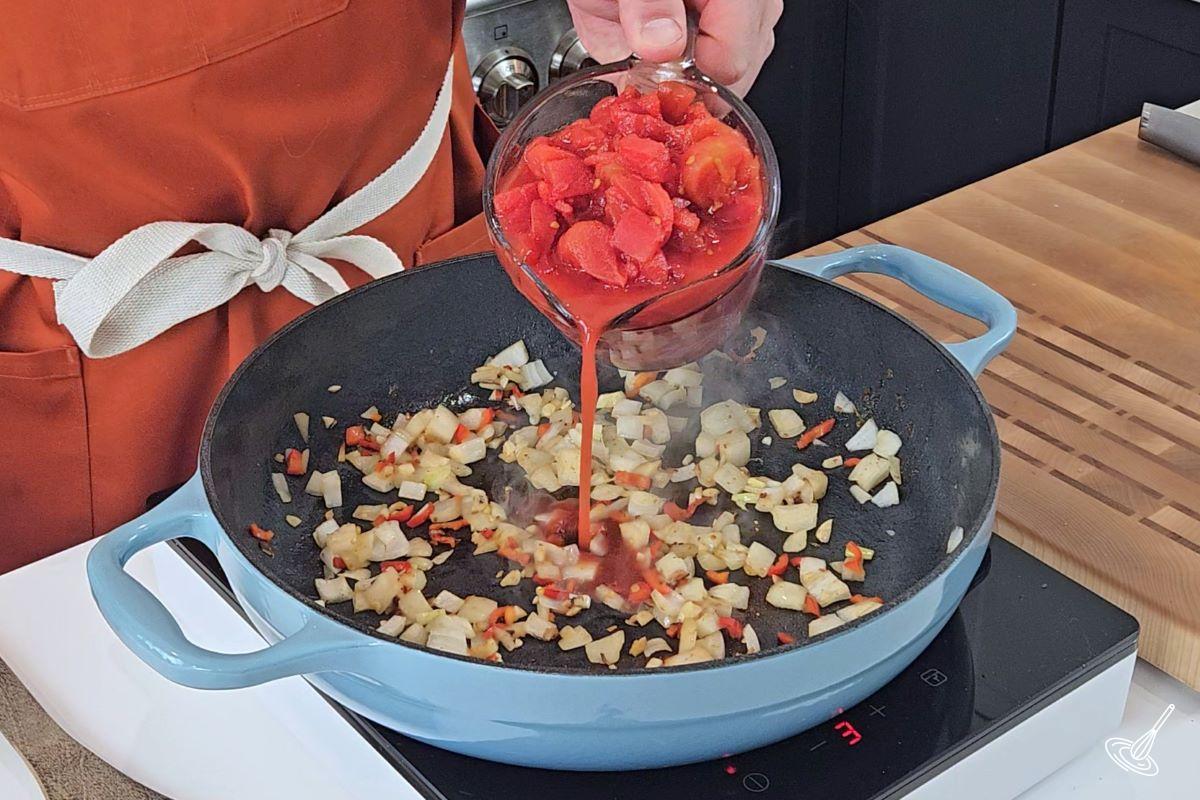Someone pouring cubed tomatoes in a skillet with onion, garlic, and peppers. 