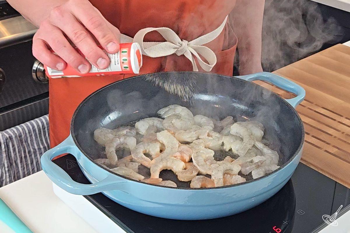 Someone sprinkling seasoning on shrimp cooking in a skillet. 