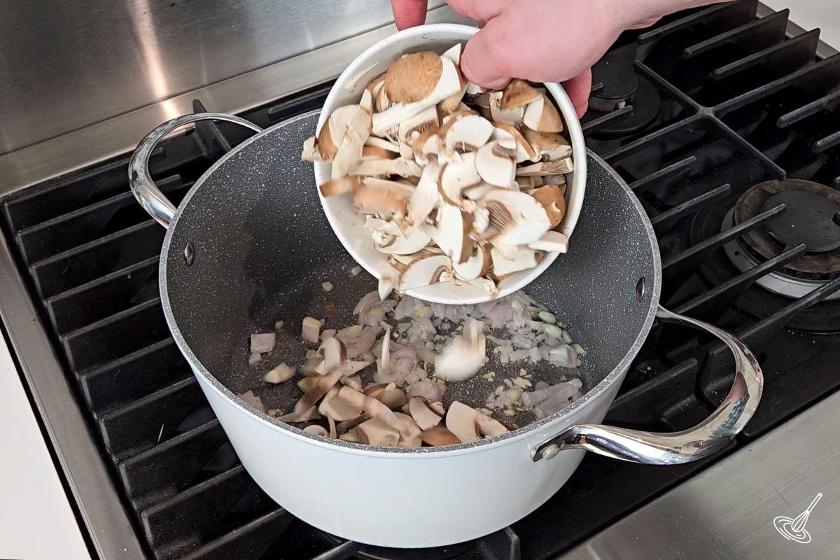Someone adding chopped mushrooms to a soup pot with shallots.
