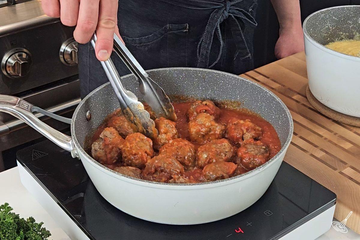 Someone adding cooked meatballs to a large skillet with tomato sauce.