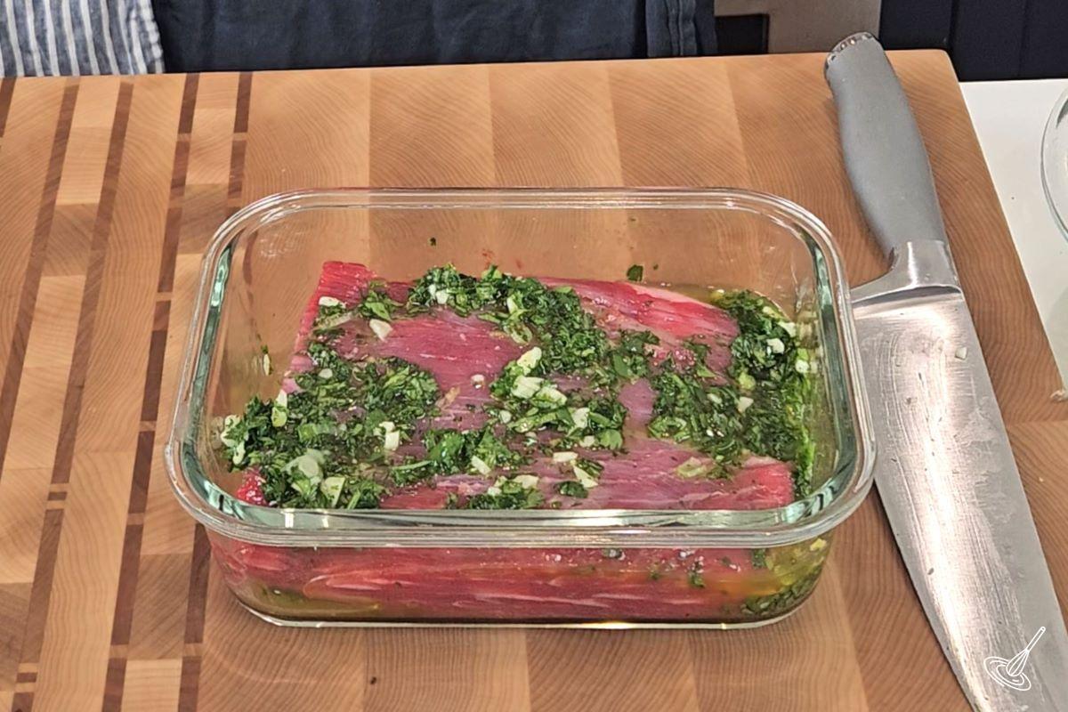 A flank steak in a container, smothered in chimichurri sauce.