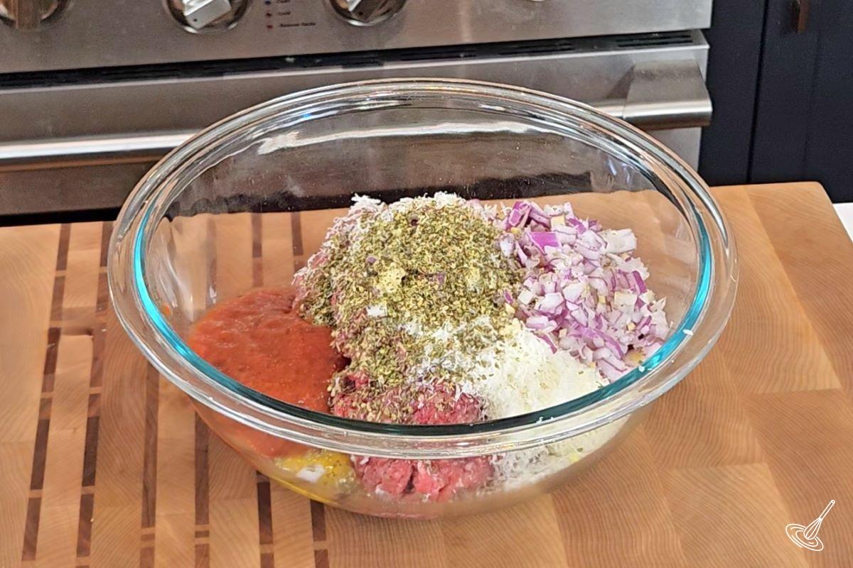 A large bowl containing ground beef, chopped onion, panko crumbs, an egg and herbs.