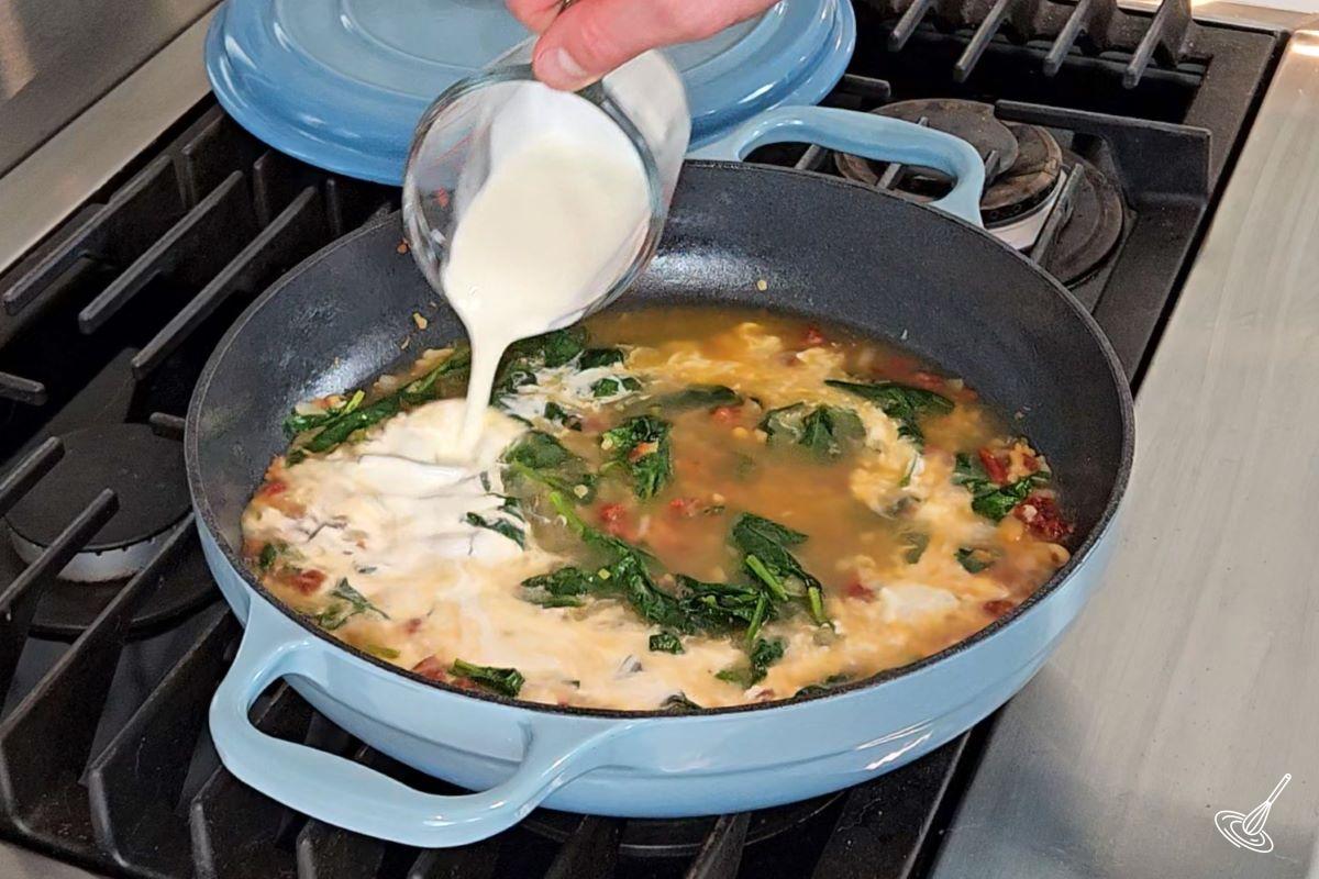 Someone pouring cream in a large skillet containing veggies and broth.