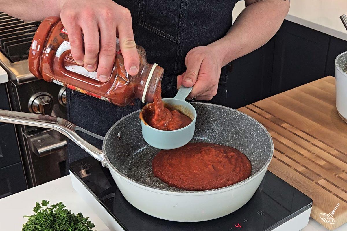 Someone adding tomato sauce to a skillet. 