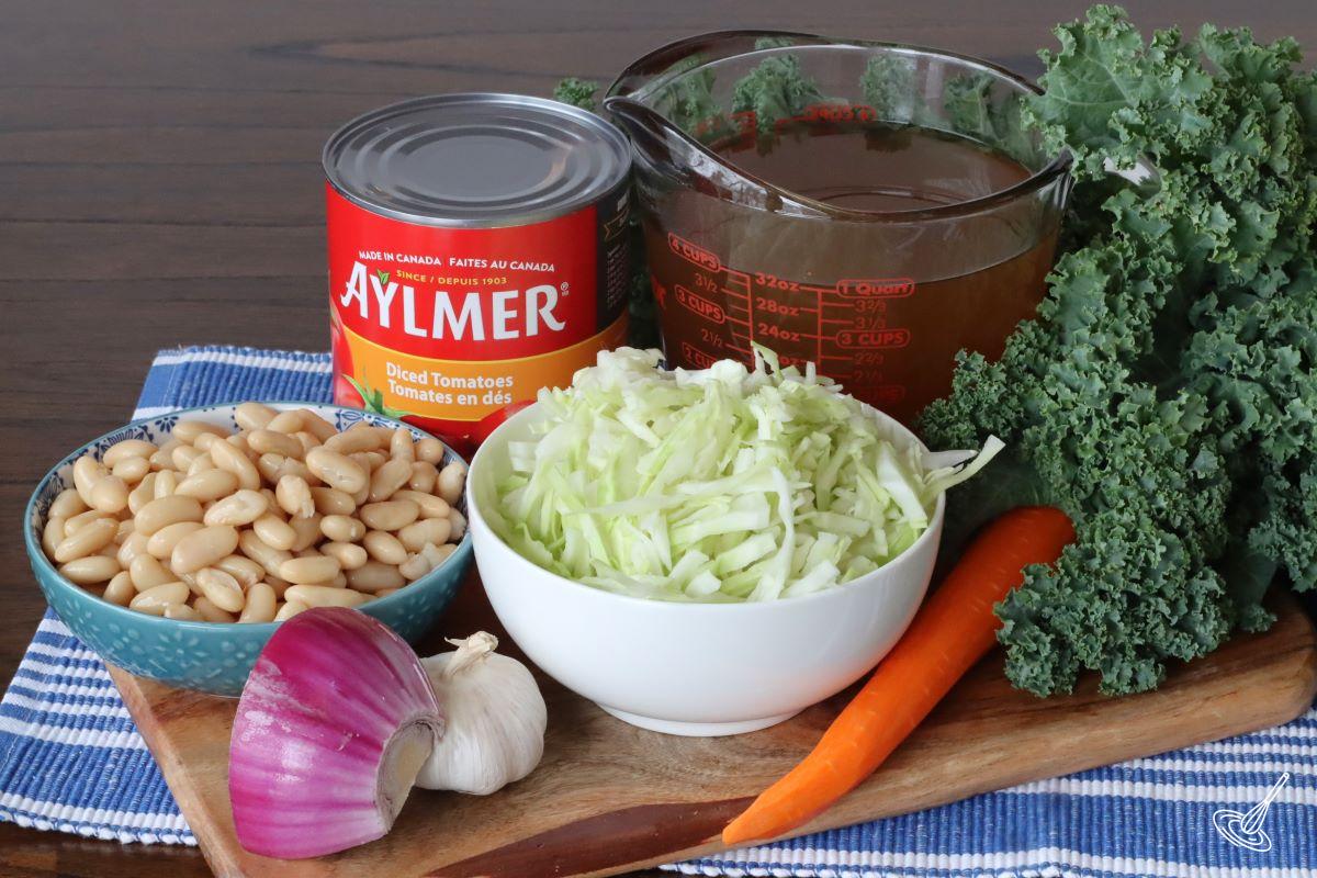 Ingredients on a board, including vegetable broth, cannelloni beans, onion, cabbage and canned tomatoes.