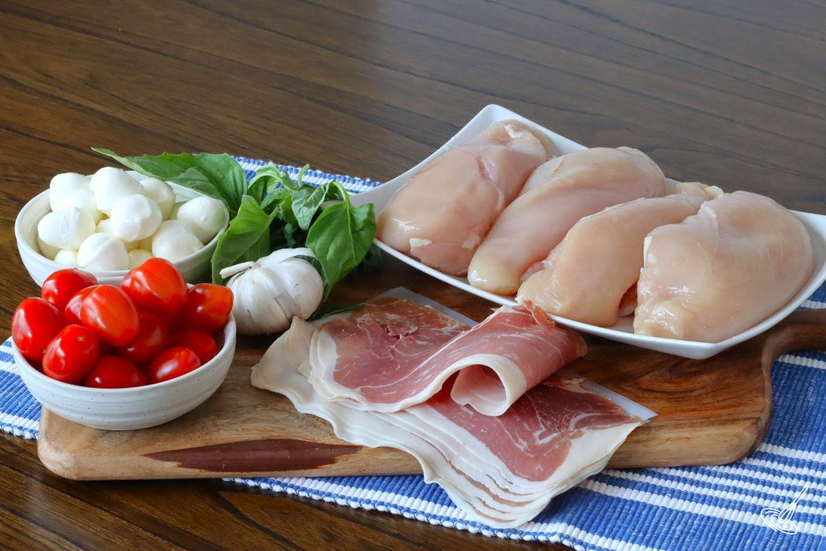Ingredients on a board, including chicken, prosciutto, tomatoes, basil, and bocconcini.