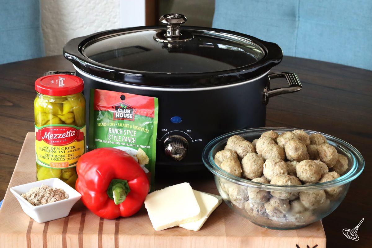 A crockpot on a table with ingredients like meatballs, peperoncini pepper, and butter.