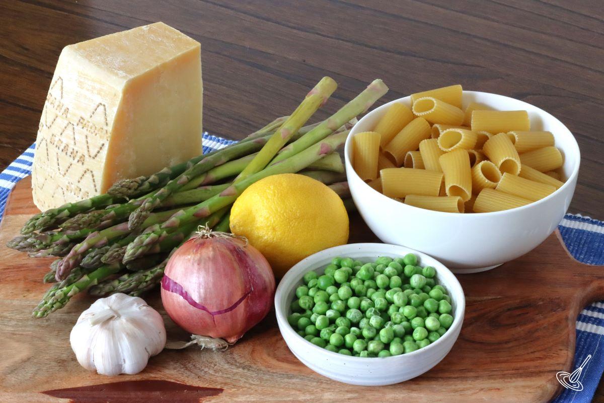 Ingredients on a wooden board, including green peas, asparagus, onion, garlic, lemon and parmesan cheese.
