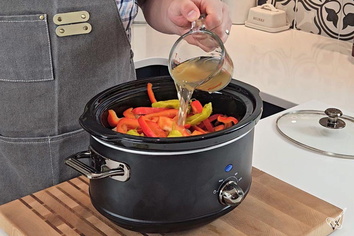 Someone pouring broth in the crock of a slow cooker with meatballs and veggies.