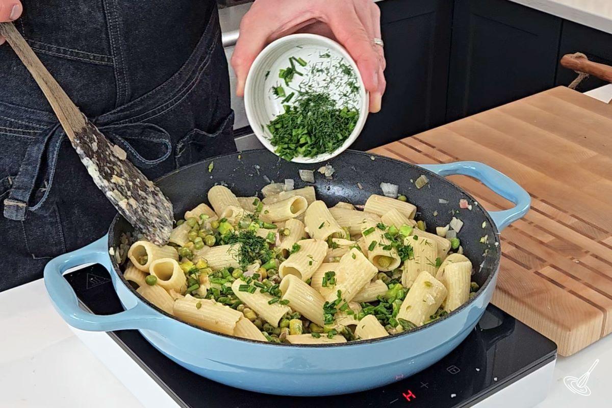 Someone sprinkling chopped dill over pasta primavera.