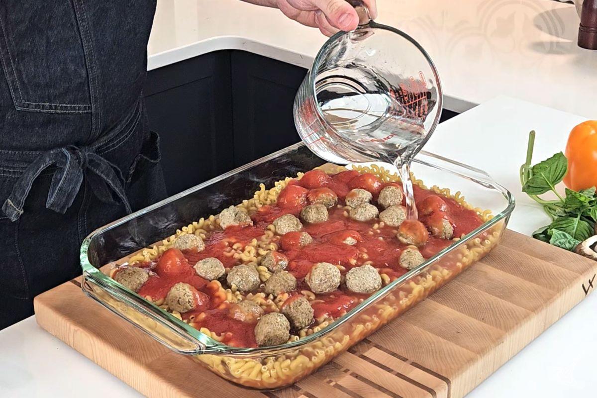 Someone pouring water into a baking dish including meatballs, pasta, and tomato sauce. 