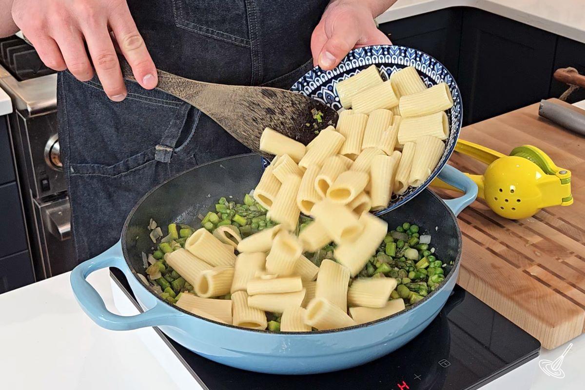 Someone adding cooked pasta to a skillet with cooked vegetables.