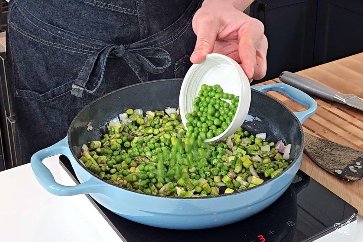 Someone adding green peas to a large skillet with asparagus onion and garlic.