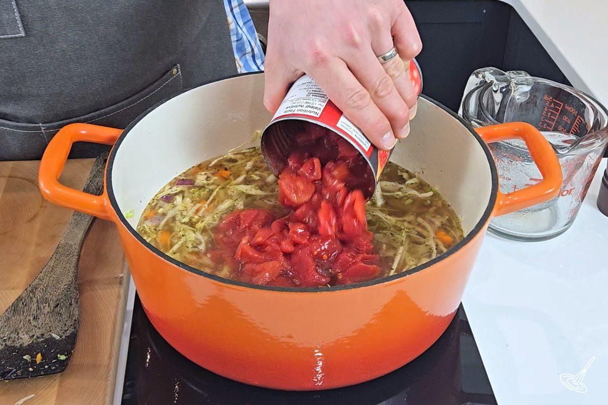 Someone pouring out a can of diced tomatoes in a Dutch oven with vegetables. 