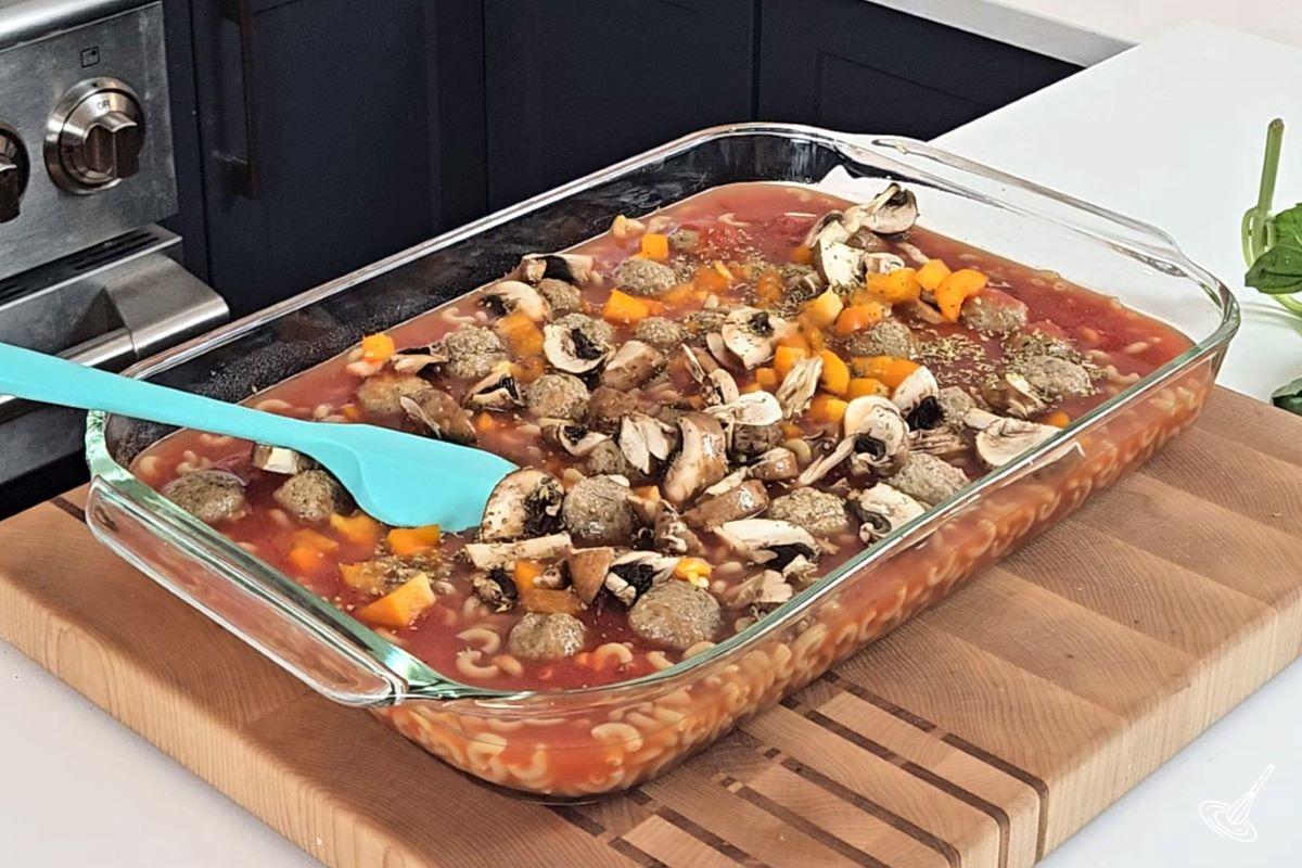 A baking dish containing pasta, tomato sauce, meatballs, chopped mushrooms, and bell pepper. 