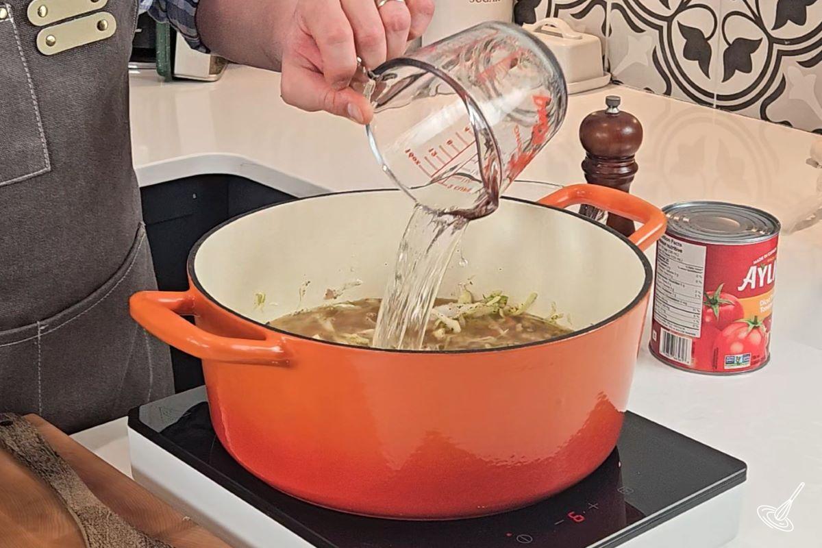 Someone pouring water in a large Dutch oven containing vegetables.