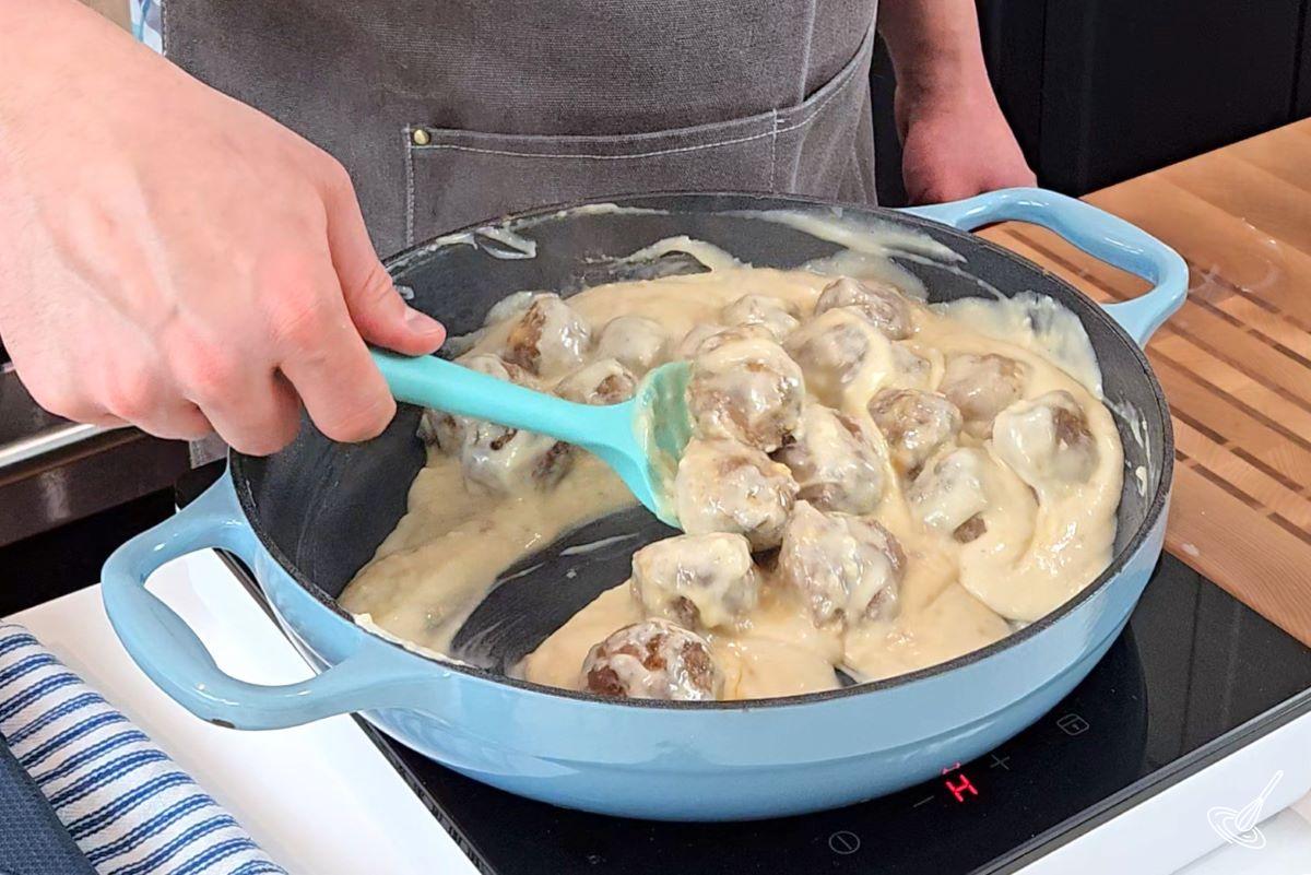 Someone stirring cooked meatballs in alfredo sauce to coat them.