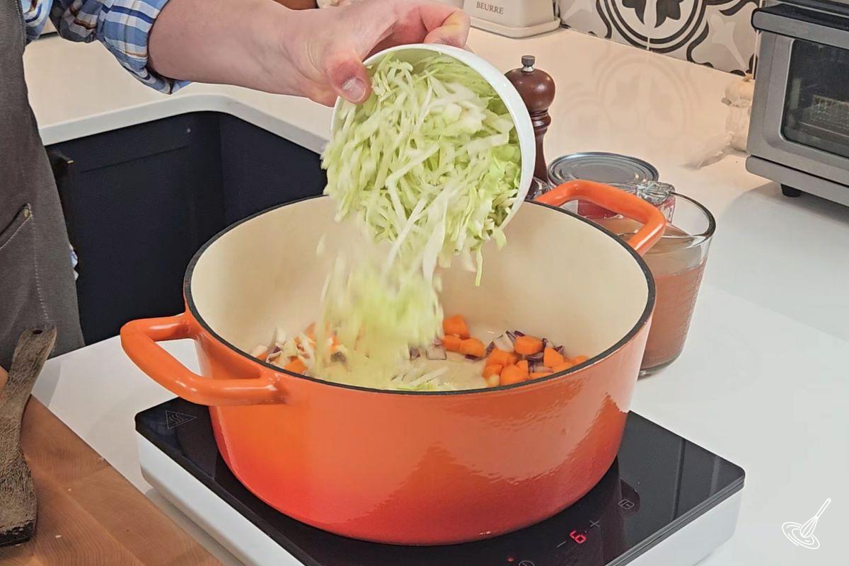 Someone placing shredded cabbage in a Dutch oven with other vegetables.