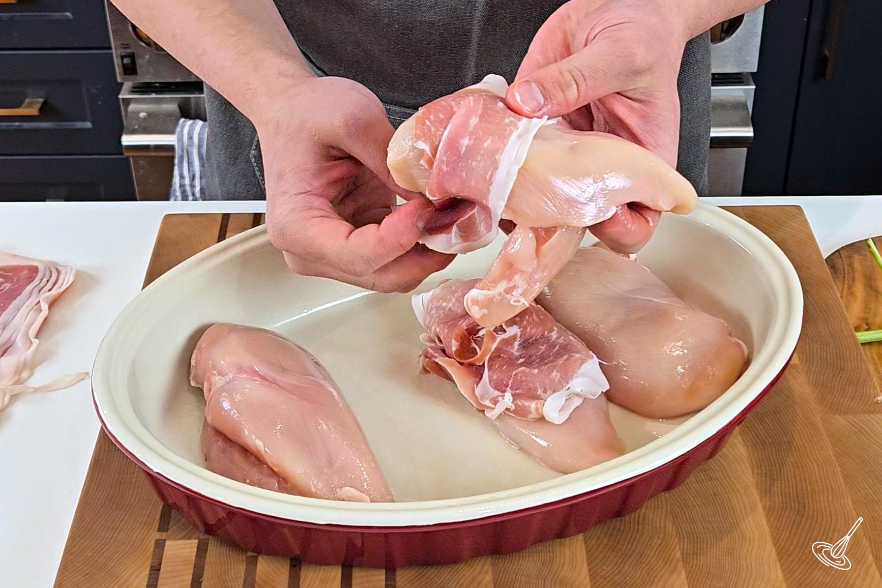Someone wrapping a chicken breast with a slice of prosciutto.