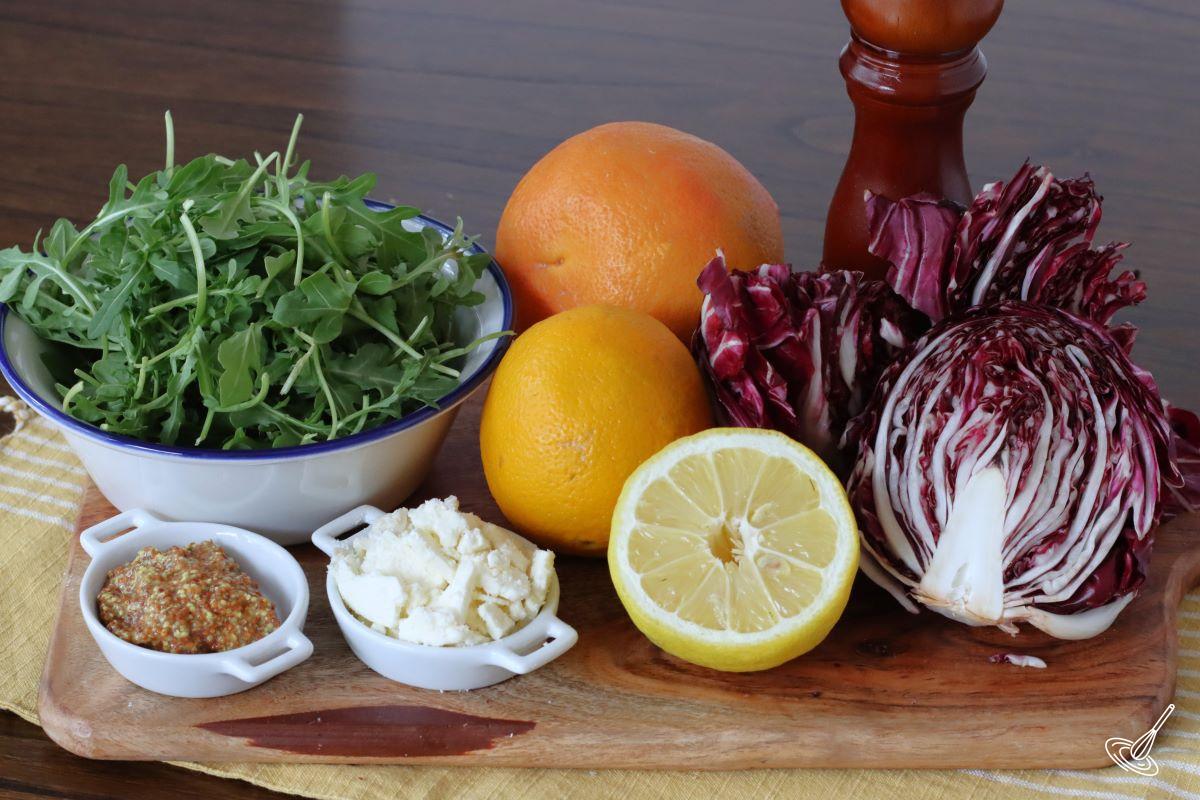 Ingredients on a wooden board including; radicchio, arugula, an orange, a grapefruit, and feta cheese.