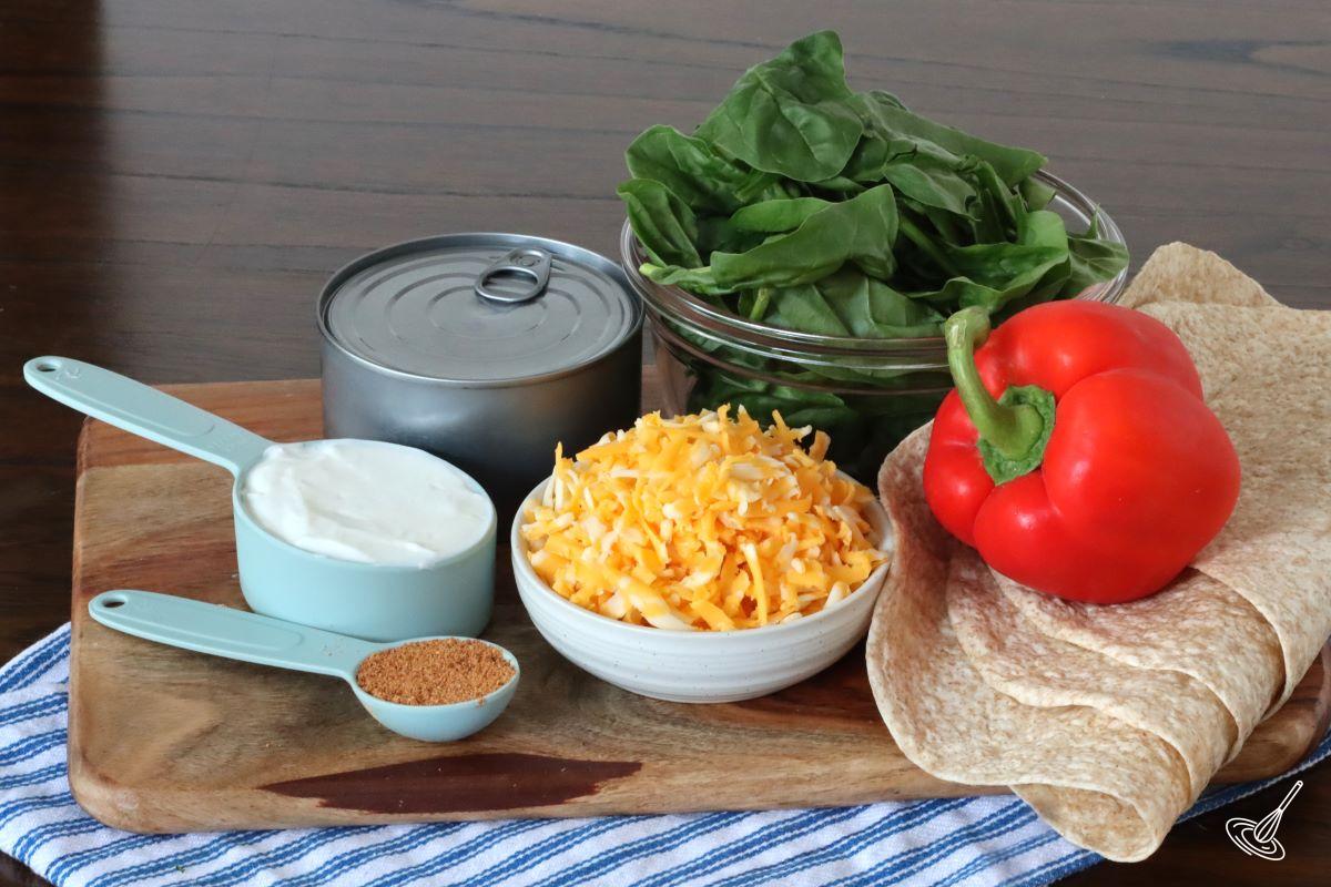 Ingredients on a wooden board including, grated cheese, canned chicken, spinach and Greek cheese.