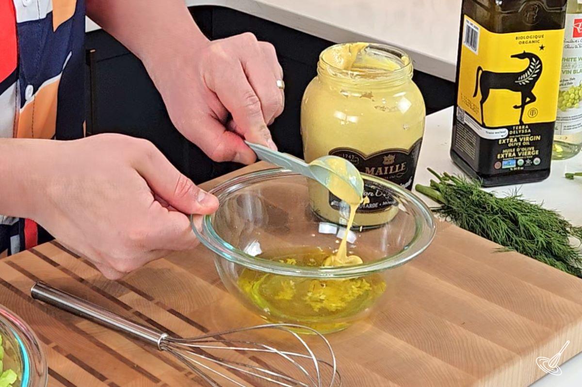Someone adding Dijon mustard to a bowl of olive oil.