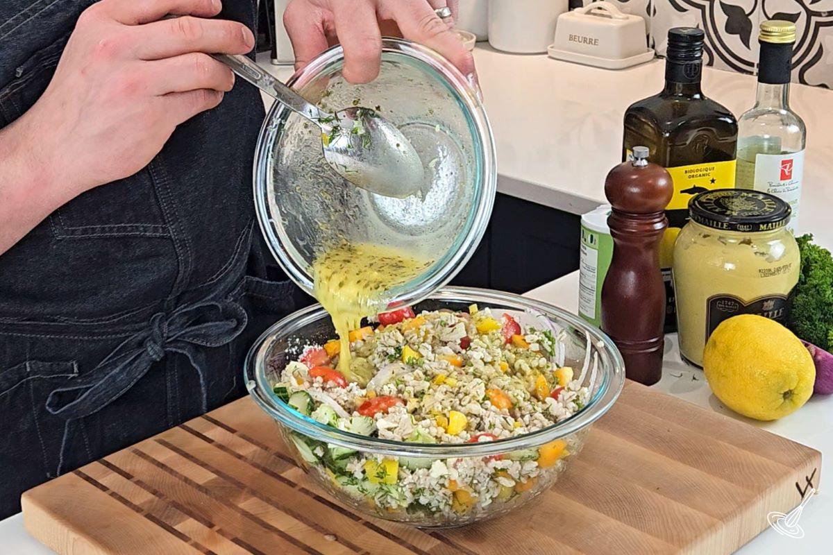 Someone pouring salad dressing over a bowl of Mediterranean Barley Salad. 