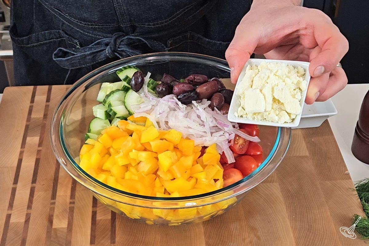 Someone adding feta to a large bowl containing vegetables. 