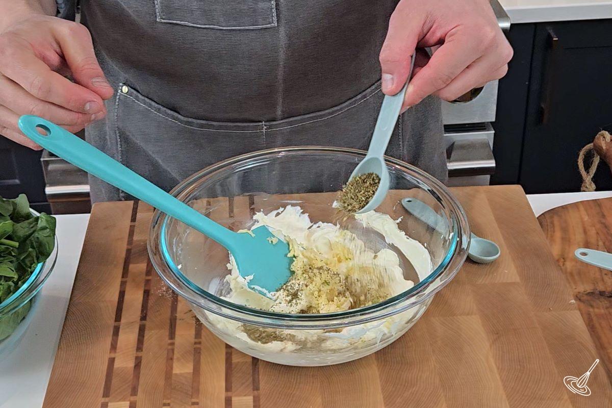 Someone adding dry oregano to a large bowl with cream cheese.