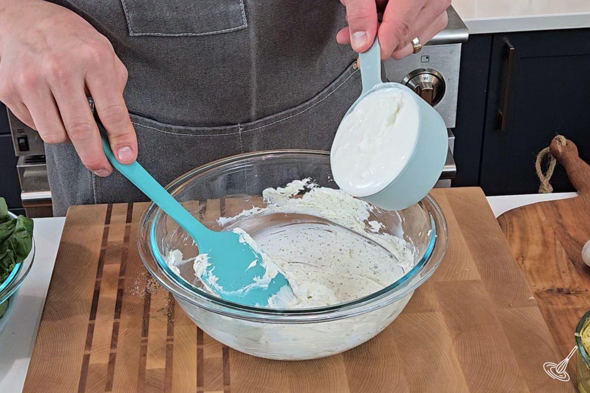 Someone adding Greek yogurt to a large bowl of softened cream cheese.