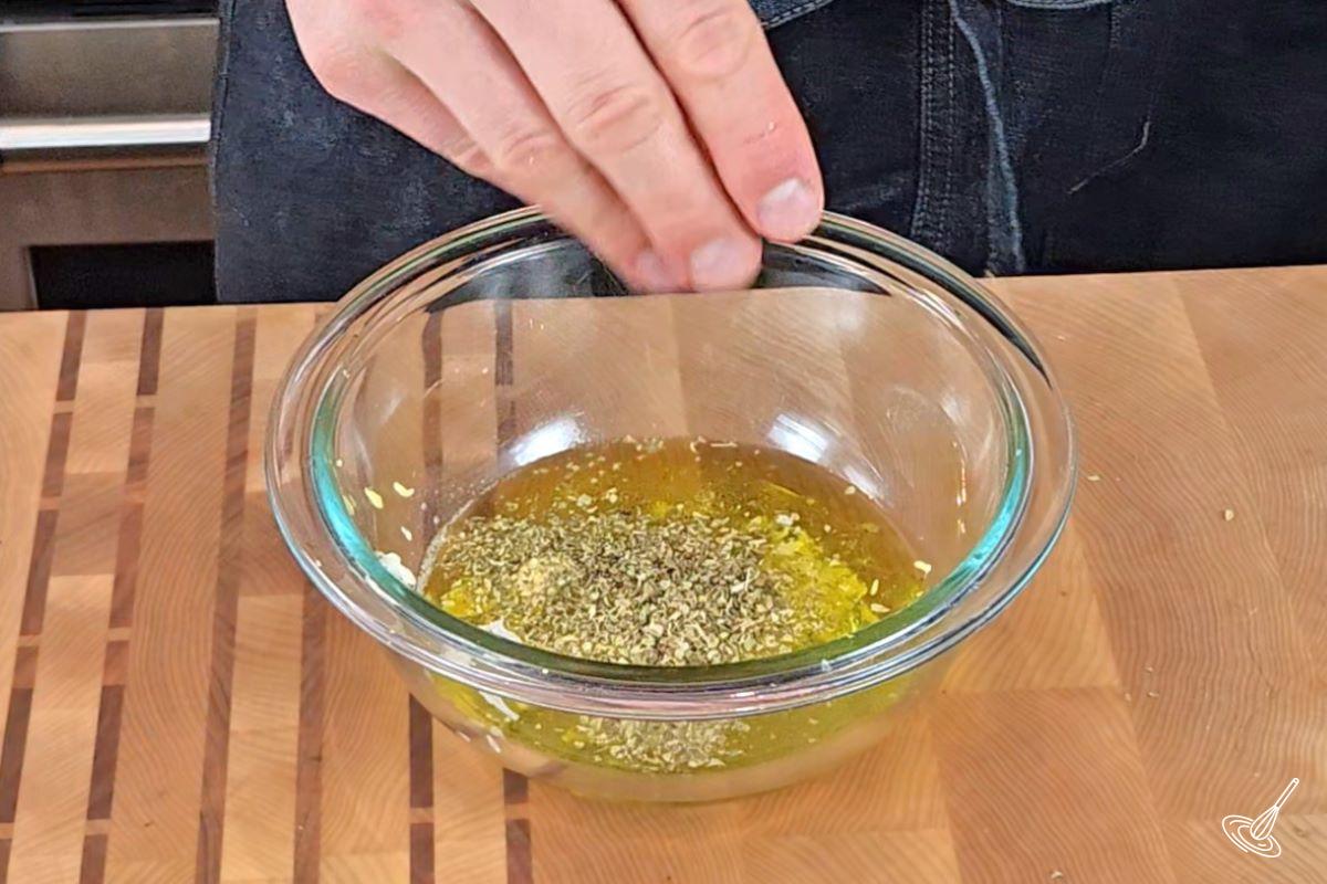 Someone adding oregano to a small bowl of salad dressing.