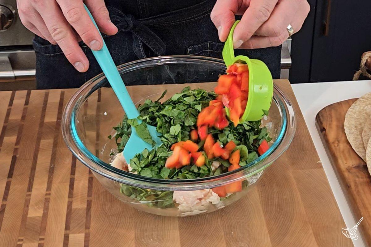 Someone adding chopped bell pepper to a large bowl with chopped spinach.