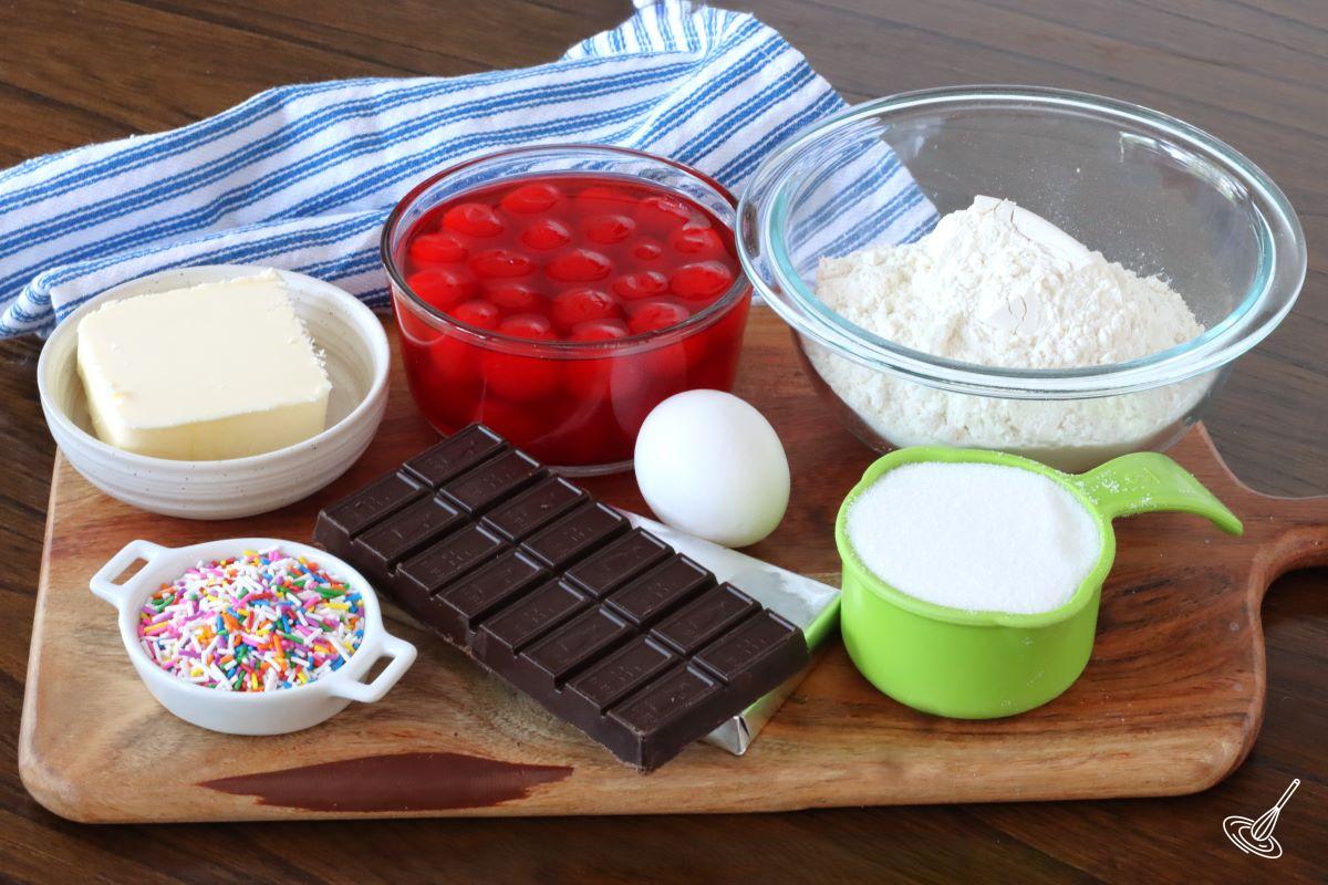 Ingredients on a wooden board including; chocolate, maraschino cherries, flour, and butter.