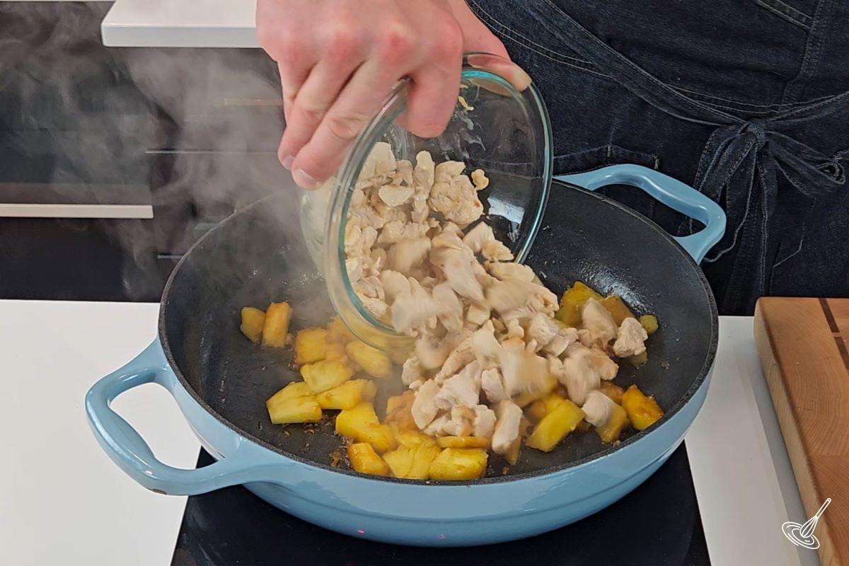 Someone adding cooked chicken to a frying pan with caramelized pieces of pineapple.