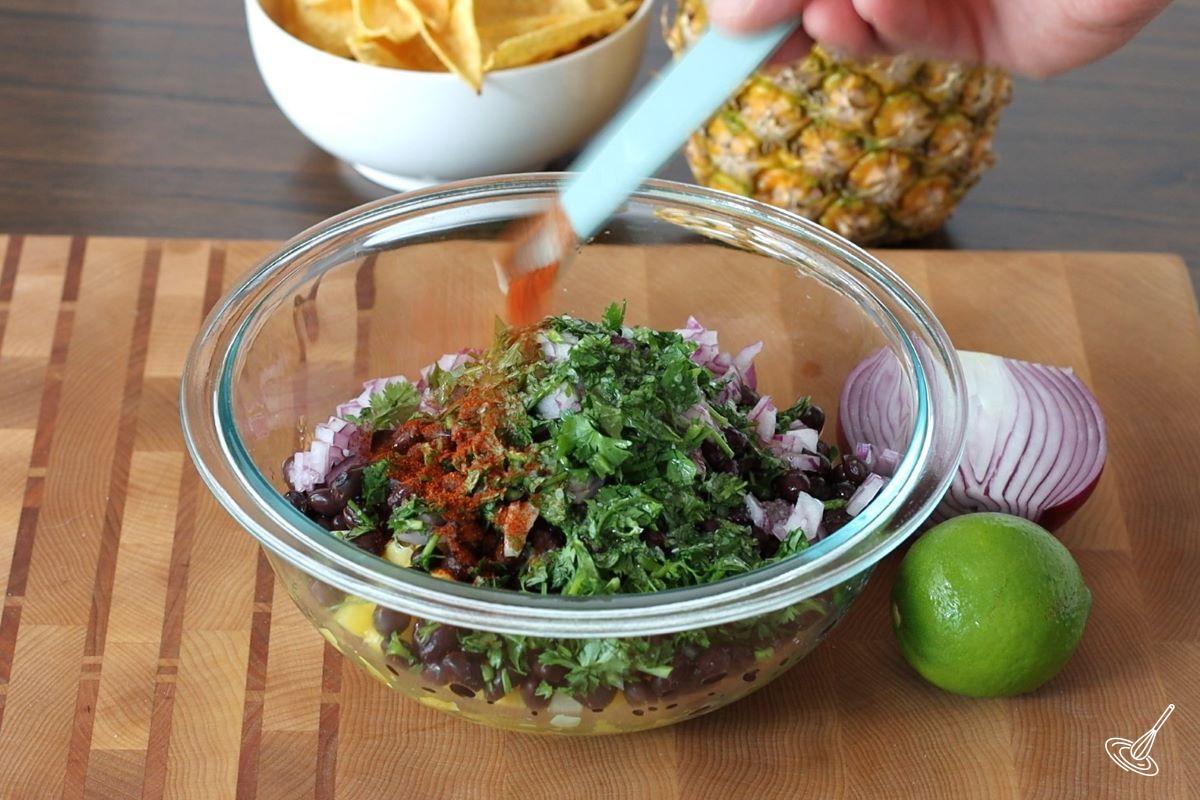 Someone adding smoked paprika to a large bowl of pineapple corn salsa.
