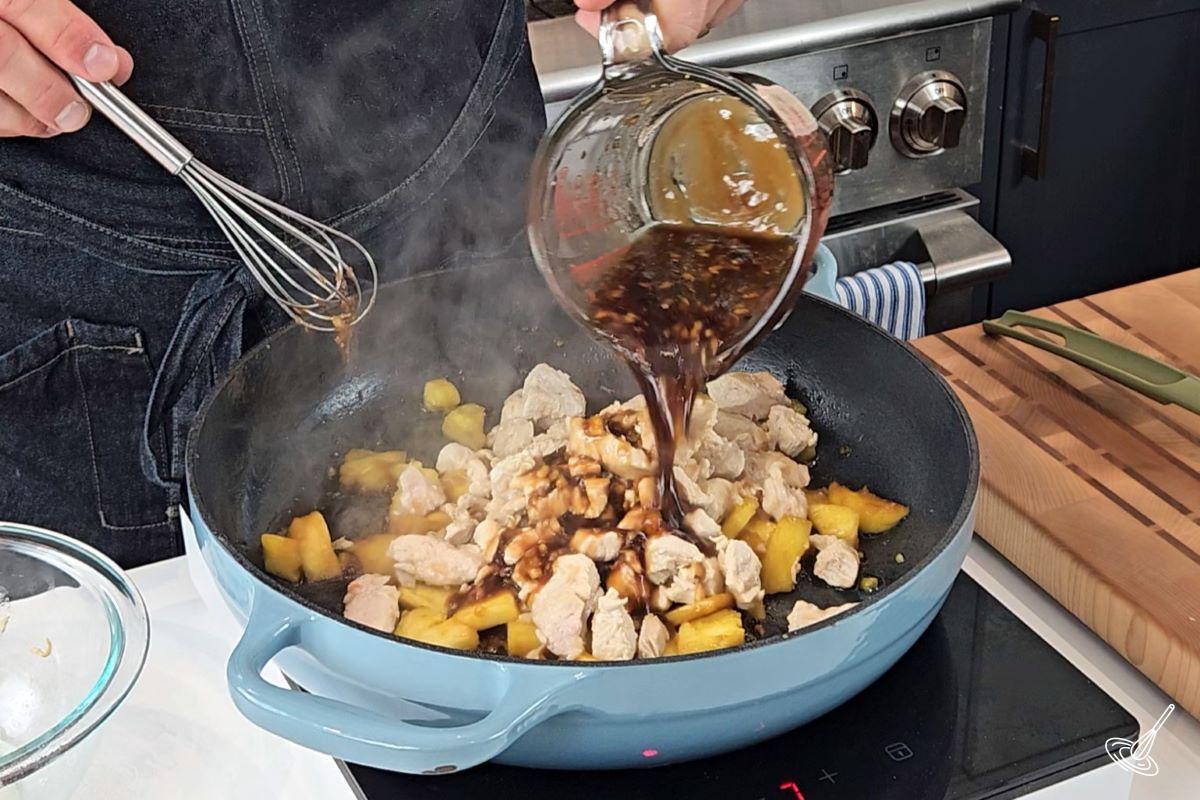 Someone pouring a soy sauce into a frying pan with chicken and pineapple