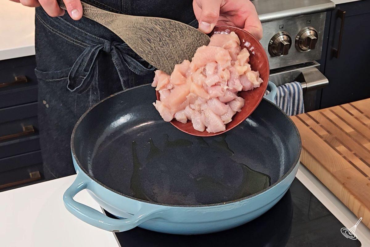 Someone places cubed chicken breasts into a large frying pan.