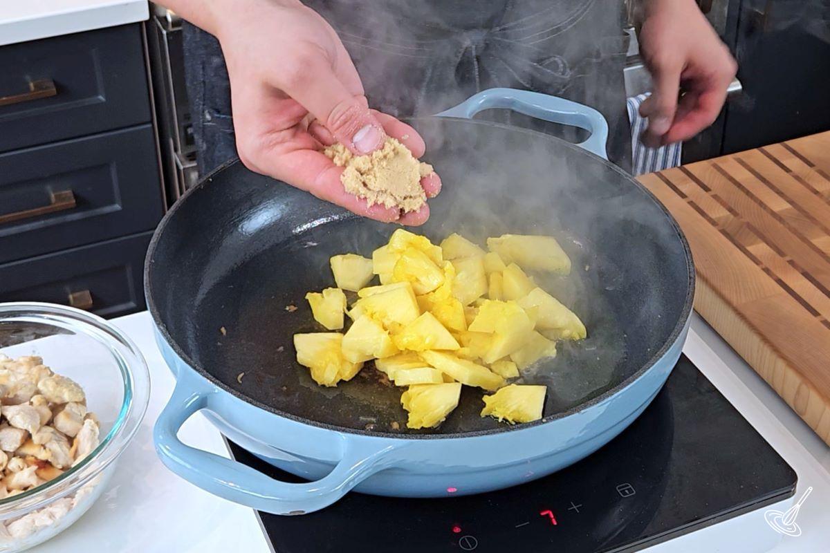 Someone adding brown sugar to a large frying pan containing chunks of pineapple.