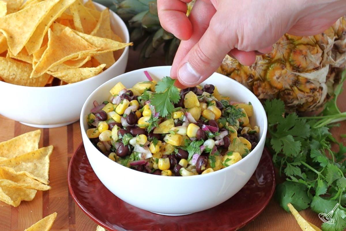 Someone placing a leaf of cilantro on top of a bowl of Pineapple Corn Salsa.