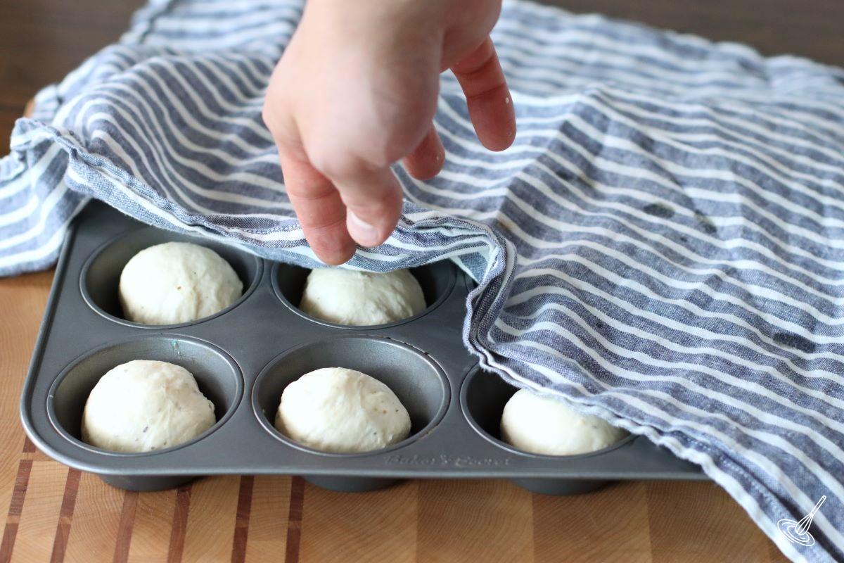 Someone placing a tea towel over balls of dough in a muffin tin. 