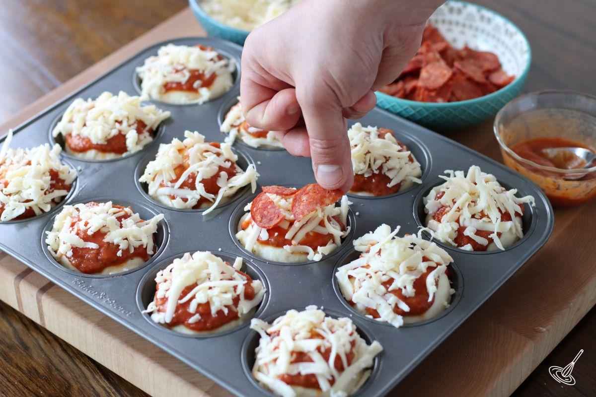 Someone placing slices of pepperoni over pizza muffins.