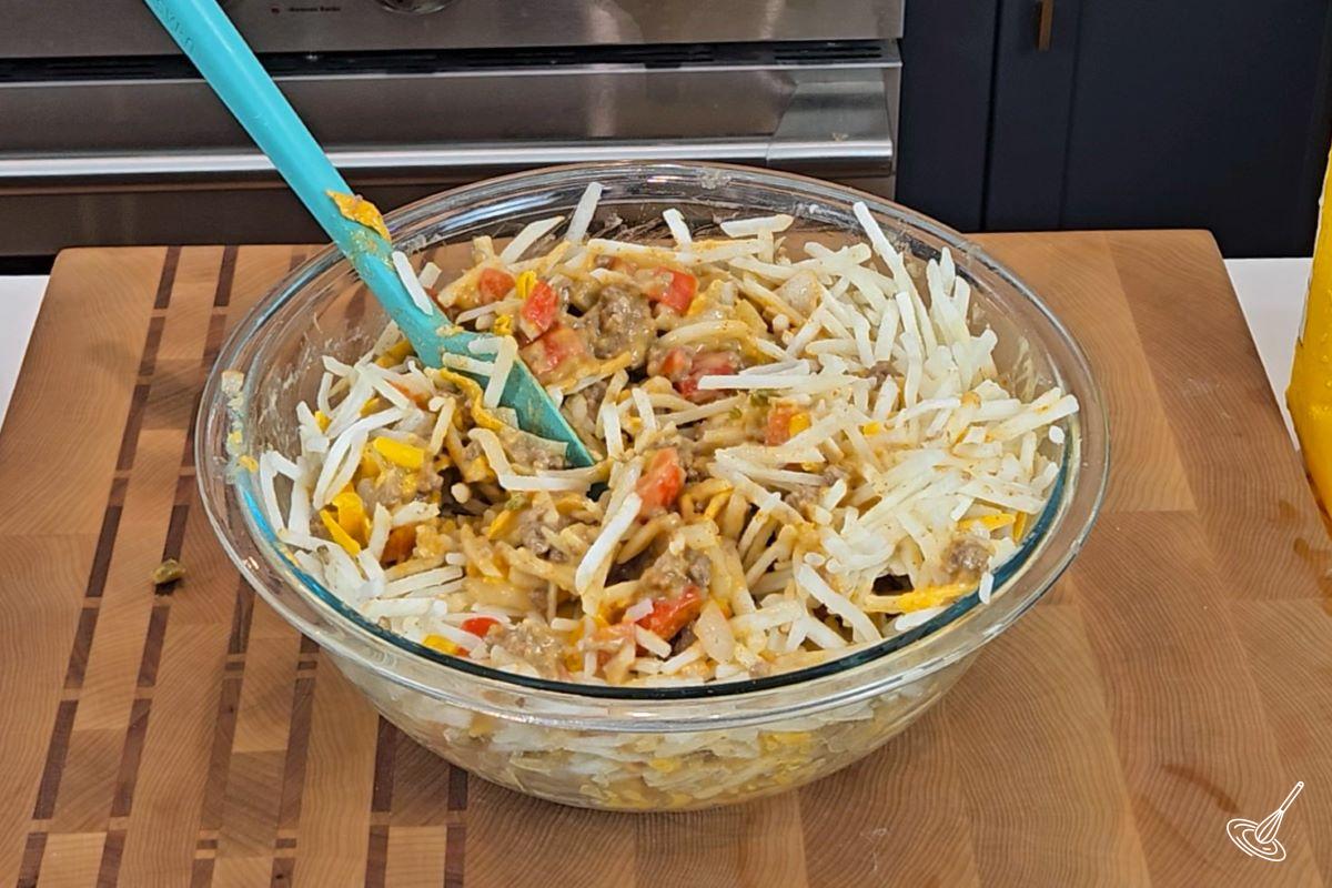 Someone stirring in frozen hashbrowns into a cooked ground beef and vegetable mixture.