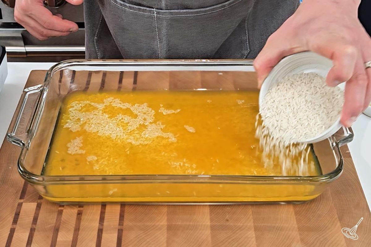 Someone adding rice to a large baking dish which contains broth. 