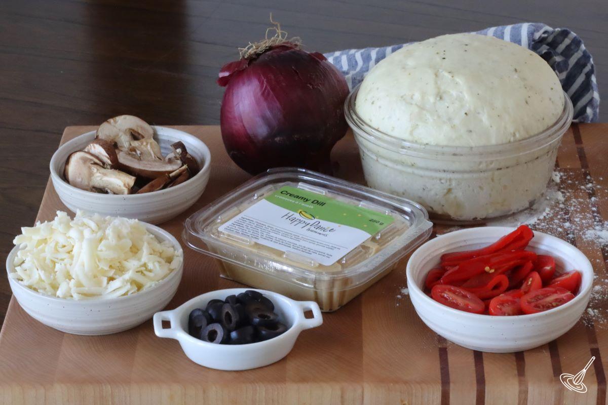 Ingredients on a cutting board including hummus, pizza dough, cheese, and vegetables. 